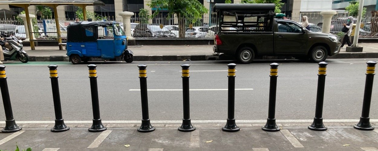 9 steel bollards across a wide pedestrian walkway as it intersects a city street.